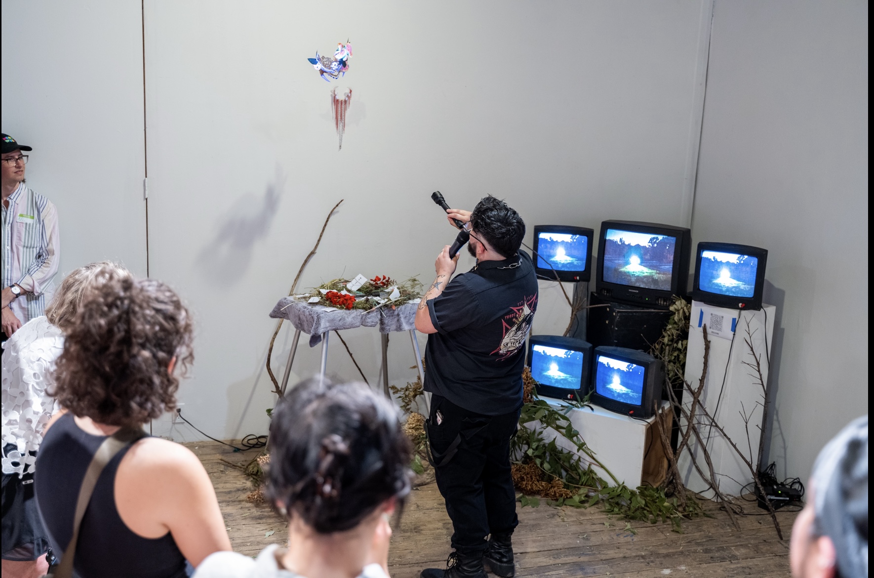 Me showing everyone the UV reactive mask with the flashlight and speaking on the mic. Behind me the video is playing and shows the field we filmed in. branches decorate the installation.