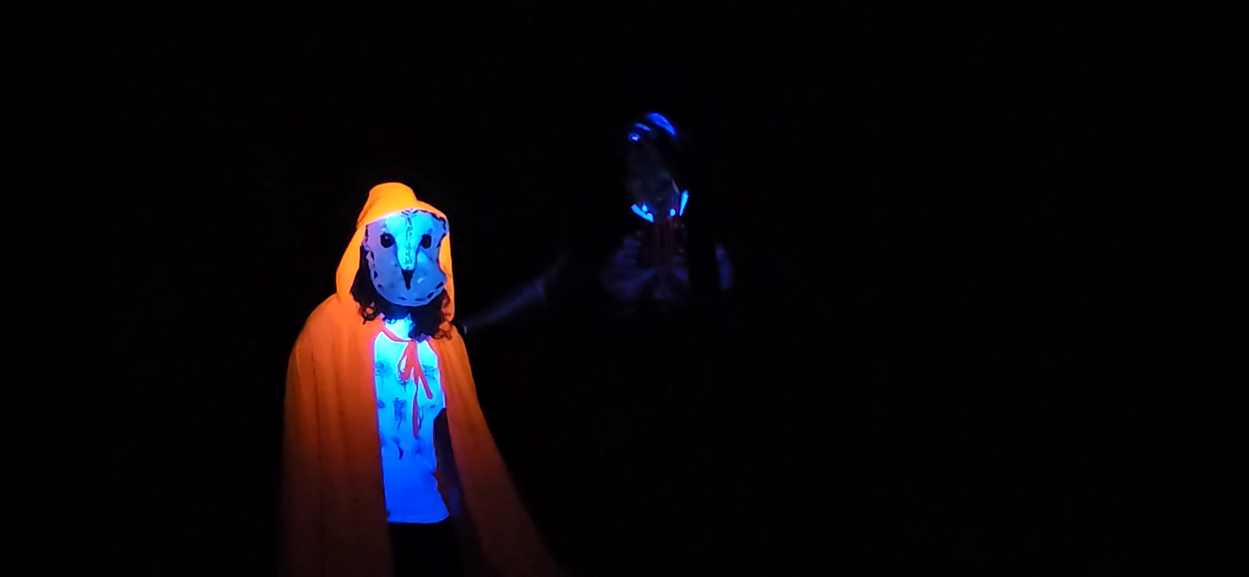 Laura and I standing in our costumes. Her cape is bright orange being lit up by the UV flashlight and shes wearing her owl mask. The only things lit up by the flashlight on my costume are the top of my white mask and the fake teeth on my mouth stretcher.