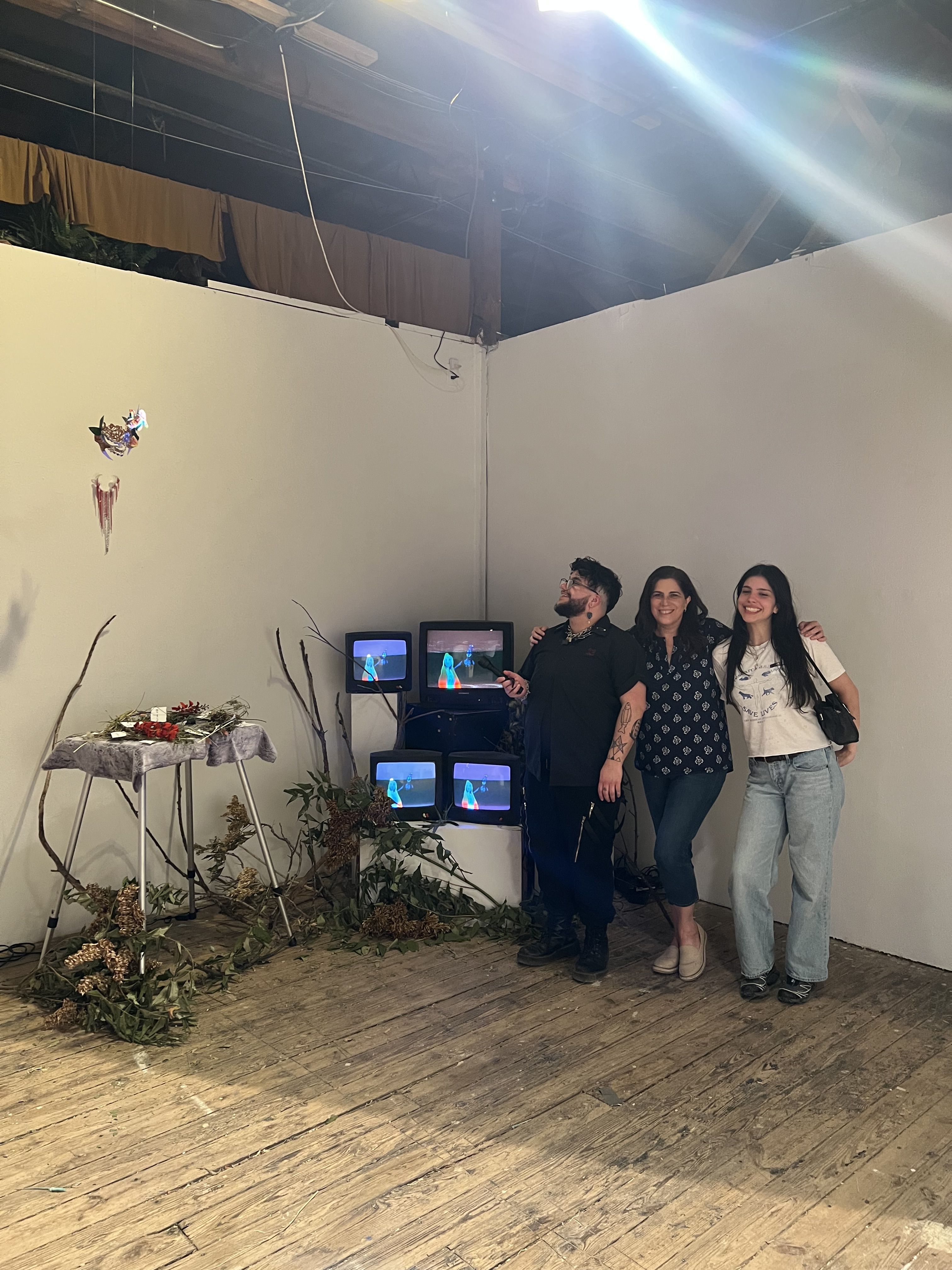 My mom, my sister and I standing in front of my installation. I am using the flashlight to illuminate the mask while the screens behind us show Laura and myself in our costumes on the video playing in the background. Branches are scattered in front of the screen and the the table to the left that holds the flashlights.