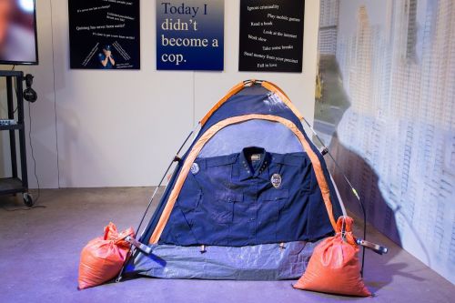 View of APD Decruitment at Fusebox. Blue and orange tent, held down by orange sandbags. Tent has a police uniform over the front. Posters in the background.