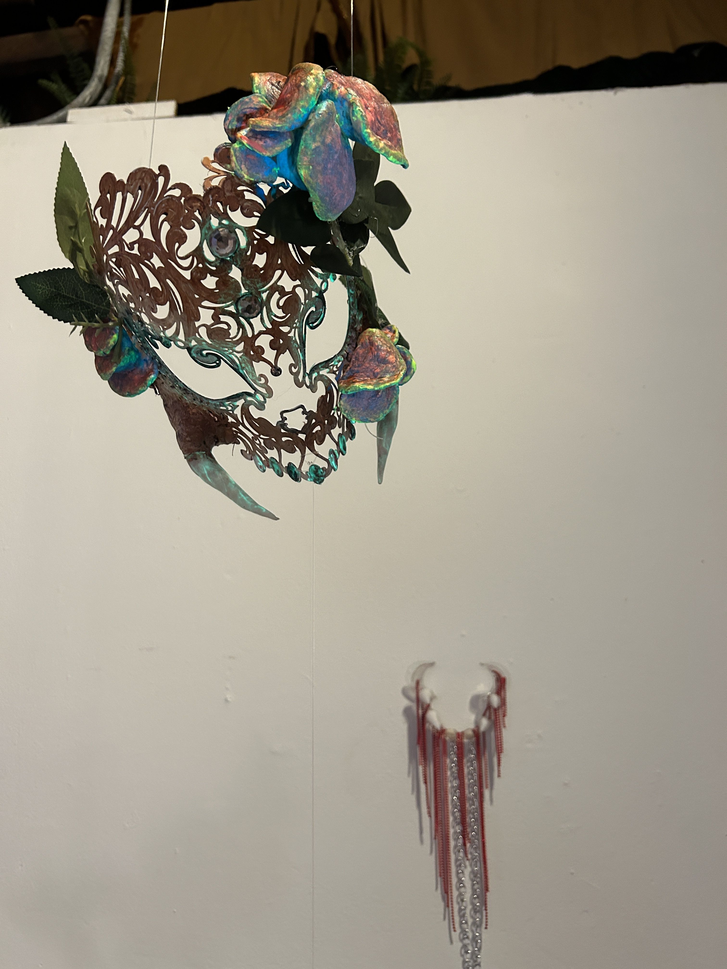 My maks hanging at MoHA. It’s a steel masks with a lace-like design cut into it. I painted it a light brown and white. On it, UV reactive green, blue, and orange colored mushrooms, plastic fangs I glued on and some fake leaves. Right under it is my mouth stretcher mounted on the wall with some red rhinestones and chains hanging from it and fake teeth glued on.